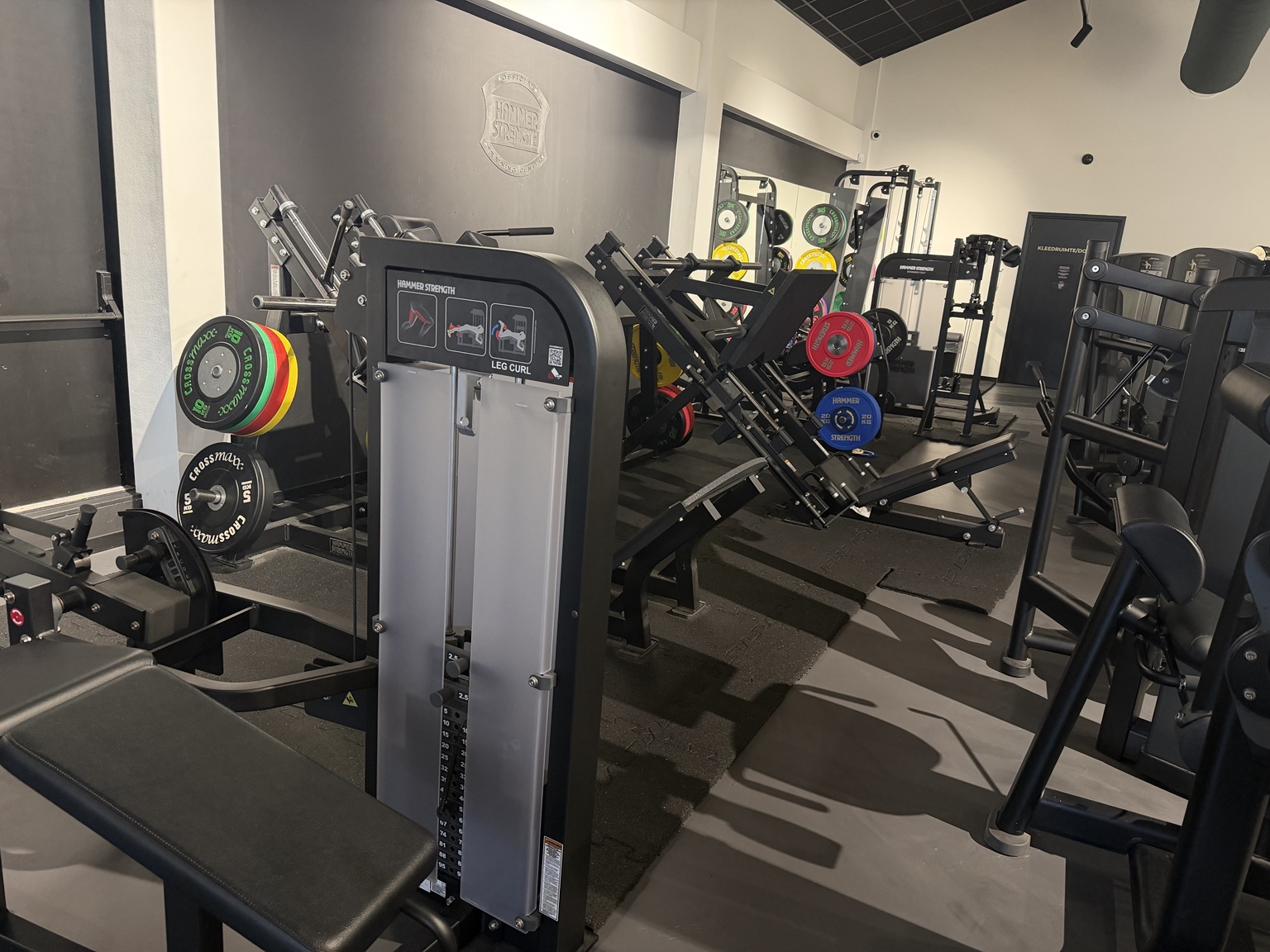 De huidige krachtvloer bij Vital-GO met Hammer Strength leg curl, plate-loaded beenapparatuur en kleurrijke halterschijven.